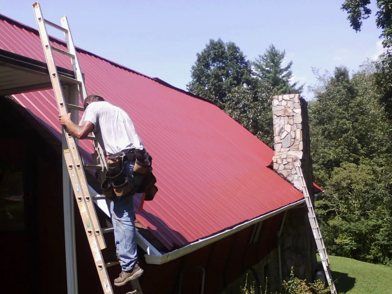 Roofer on a ladder beside a red metal roof and stone chimney — Soffit & Fascia Repair by Roofing Brothers in Long Prairie